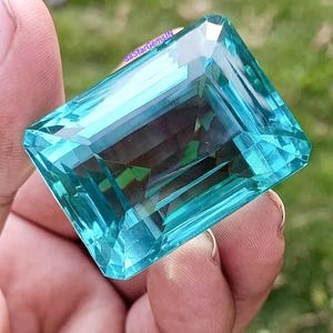 May include: A large, rectangular, aquamarine gemstone with multiple facets. The gem is a vibrant, transparent blue-green color, reflecting light and creating a sparkling effect. The image includes the text "SaStarGemsIN".