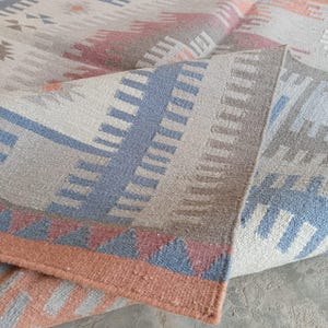 Könnte beinhalten: Ein gewebter Teppich mit einem geometrischen Muster in gedeckten Tönen von Altrosa, Schieferblau und Creme. Das Design besteht aus sich wiederholenden Dreiecken, vertikalen Streifen und Treppenmotiven, wobei eine umgeklappte Ecke die Textur zeigt.