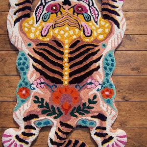 Peut inclure: Un tapis touffeté à la main en forme de tigre. Il présente un design détaillé avec un visage jaune, des rayures noires et des motifs floraux roses, oranges et bleus. Le tapis est posé sur un sol en bois.