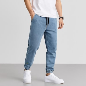 May include: Light blue denim joggers with a drawstring waist and elasticated cuffs. The trousers have a relaxed fit and are worn with white trainers. A small detail of yellow and blue stitching is visible on the left leg.