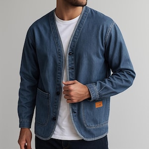 Könnte beinhalten: Blaue Jeansjacke mit V-Ausschnitt, Knopfverschluss und zwei Vordertaschen. Die Jacke hat ein kleines orangefarbenes Etikett auf der Vorderseite. Das Model trägt ein weißes T-Shirt darunter.