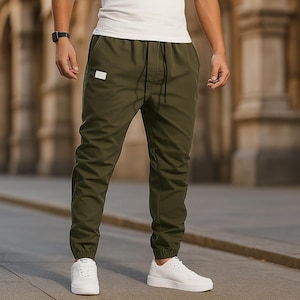 May include: Olive green joggers with a drawstring waist and elastic cuffs. The pants are paired with white sneakers and a white t-shirt. A small white tag is visible on the left leg. The background features a blurred outdoor setting.