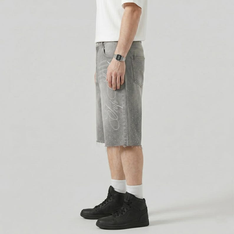 May include: Gray denim shorts with a raw hem and a white script design on the side. The shorts have a faded appearance and are paired with black high-top sneakers.