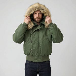 K&ouml;nnte beinhalten: Olivgr&uuml;ne Bomberjacke mit Kapuze mit Kunstfellfutter. Die Jacke hat eine Knopfleiste, gerippte B&uuml;ndchen und einen gerippten Saum. Das Model tr&auml;gt eine dunkelblaue Jeans. Ein klassisches Oberbekleidungsst&uuml;ck.
