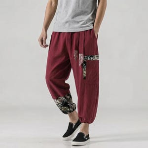 May include: Burgundy linen trousers with decorative patches and cuffs. The trousers have a relaxed fit and drop-crotch design. The cuffs and pocket details feature a patterned design in black, white, and gold.