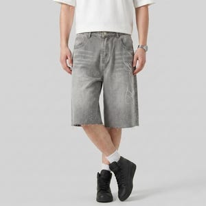 May include: Gray denim shorts with a raw hem and white decorative design. The shorts have a faded appearance and are knee-length. The model is wearing black high-top sneakers and a white t-shirt.