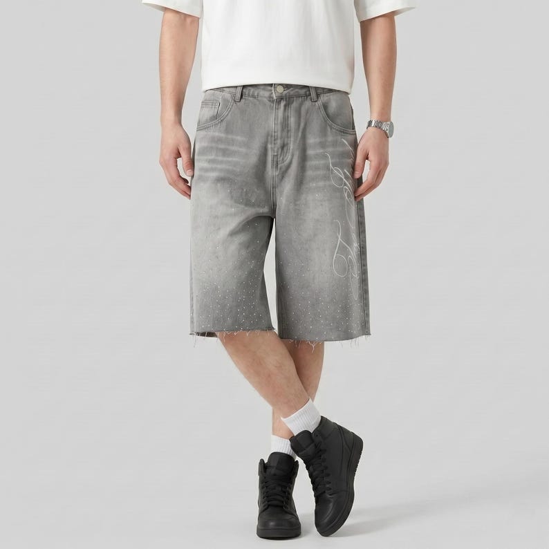 May include: Gray denim shorts with a raw hem and white decorative design. The shorts have a faded appearance and are knee-length. The model is wearing black high-top sneakers and a white t-shirt.