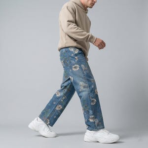 May include: Light blue denim jeans with a floral pattern. The jeans have a straight leg cut and are adorned with white and beige flowers. The person in the image is wearing a beige hoodie and white sneakers.