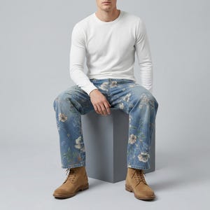 May include: A person wearing a white long-sleeve shirt, blue jeans with a floral pattern, and tan boots. The jeans feature a light blue wash with a detailed floral design in shades of white, beige, and green.