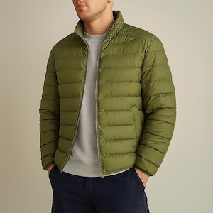 May include: Olive green puffer jacket with a stand-up collar and a full-length zipper. The jacket has horizontal stitching and a pocket on the front. The person is wearing a gray crew neck shirt underneath and navy blue pants.