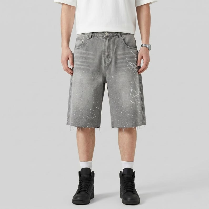 May include: Gray denim shorts with a raw hem and white splatter paint effect. The shorts feature a white decorative design on the right leg. The model is wearing black high-top sneakers and a white t-shirt.