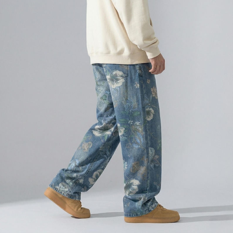 May include: Blue denim jeans with a floral pattern in white, beige, and green. The jeans have a relaxed fit and are worn with tan sneakers. The floral design covers the entire surface of the jeans.