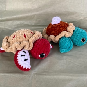 May include: Two crocheted turtle plush toys. One is tan with red detailing, resembling an apple pie. The other is teal with a brown top, resembling a pumpkin pie. Both have black and silver eyes, on a light green surface.