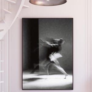 May include: Monochrome framed print of a dancer in mid-movement. The image captures the dancer's blurred form in a dark outfit and white tutu. The artwork is displayed on a white wall, creating a striking contrast.