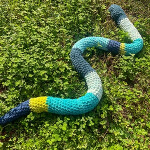 Puede incluir: Un juguete de serpiente de ganchillo en tonos de azul, verde y amarillo, sobre hierba y tr&eacute;bol verdes. La serpiente est&aacute; hecha de hilo suave y tiene un dise&ntilde;o segmentado con colores variados. El juguete est&aacute; dise&ntilde;ado para parecerse a una serpiente.