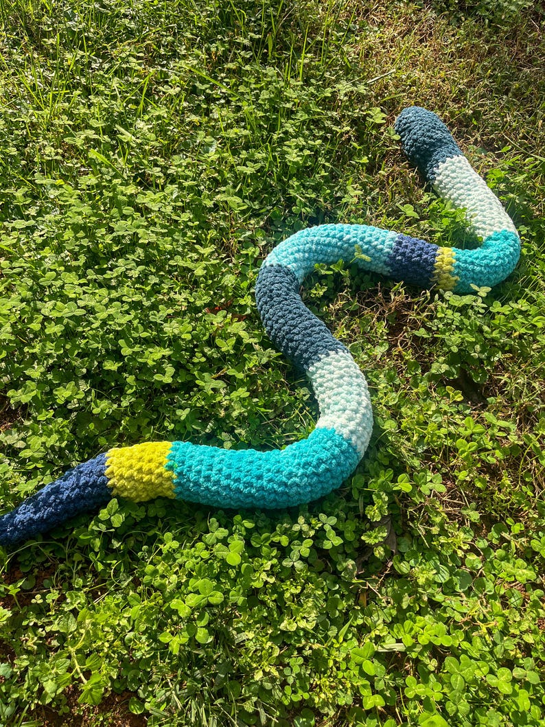 Puede incluir: Un juguete de serpiente de ganchillo en tonos de azul, verde y amarillo, sobre hierba y tr&eacute;bol verdes. La serpiente est&aacute; hecha de hilo suave y tiene un dise&ntilde;o segmentado con colores variados. El juguete est&aacute; dise&ntilde;ado para parecerse a una serpiente.