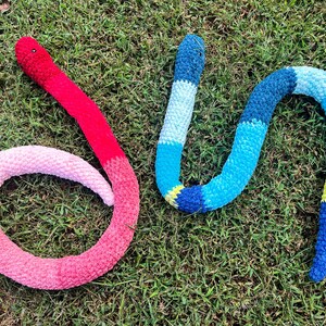 Puede incluir: Dos serpientes de ganchillo sobre hierba. Una es roja y rosa, la otra azul y amarilla. La serpiente roja tiene la cabeza roja y la cola rosa. La serpiente azul tiene la cabeza azul y la cola amarilla.