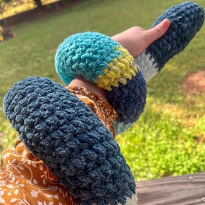 Puede incluir: Un juguete de serpiente de crochet hecho a mano en tonos de azul, turquesa, amarillo y blanco. La serpiente suave y texturizada est&aacute; enrollada alrededor de una mu&ntilde;eca. El juguete tiene una cabeza redondeada y un cuerpo largo y segmentado.