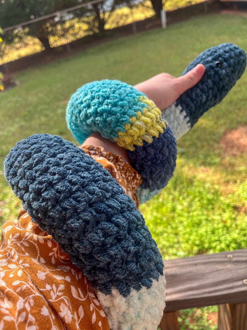 Puede incluir: Un juguete de serpiente de crochet hecho a mano en tonos de azul, turquesa, amarillo y blanco. La serpiente suave y texturizada est&aacute; enrollada alrededor de una mu&ntilde;eca. El juguete tiene una cabeza redondeada y un cuerpo largo y segmentado.