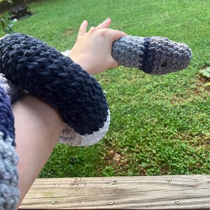 Puede incluir: Una serpiente de ganchillo hecha a mano en tonos grises, azules y blancos. El juguete de peluche tiene un cuerpo redondeado y un ojo peque&ntilde;o y oscuro. La serpiente se sostiene sobre un fondo de hierba verde y una barandilla de madera.