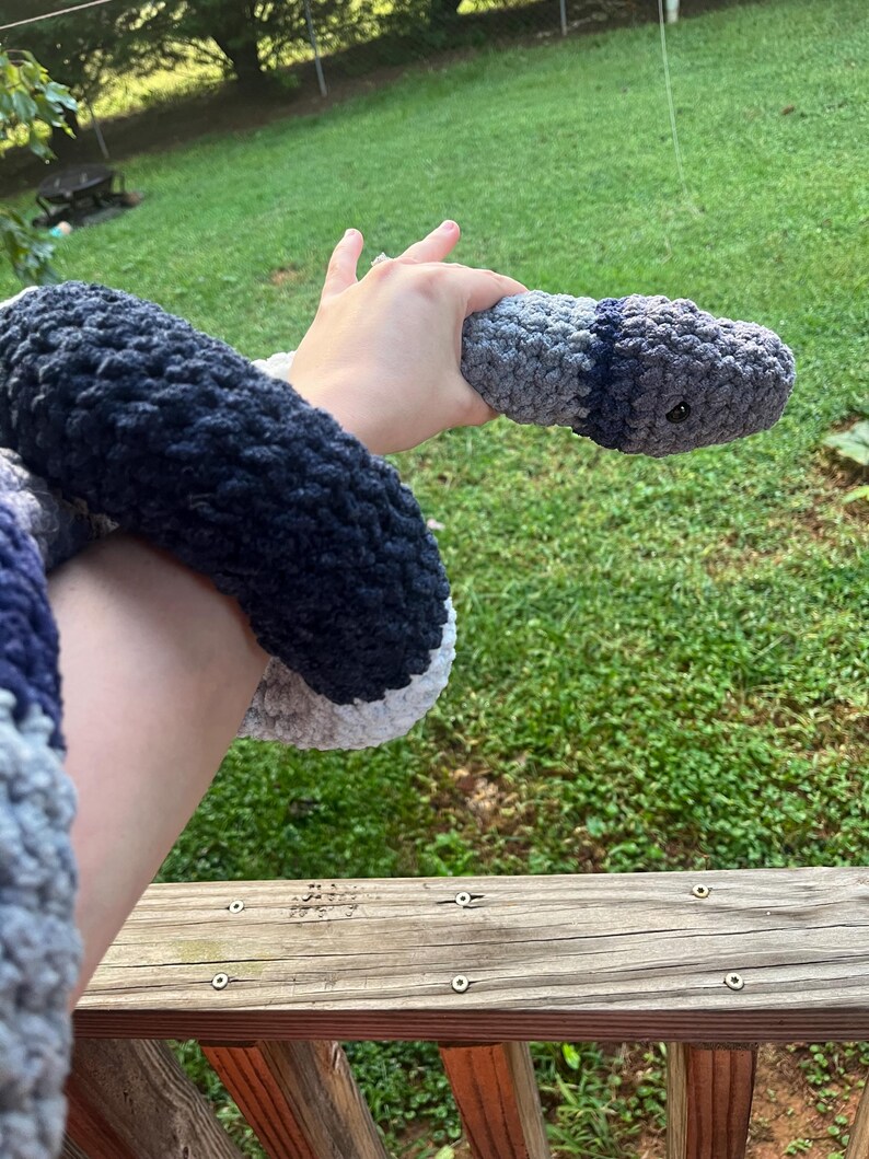 Puede incluir: Una serpiente de ganchillo hecha a mano en tonos grises, azules y blancos. El juguete de peluche tiene un cuerpo redondeado y un ojo peque&ntilde;o y oscuro. La serpiente se sostiene sobre un fondo de hierba verde y una barandilla de madera.