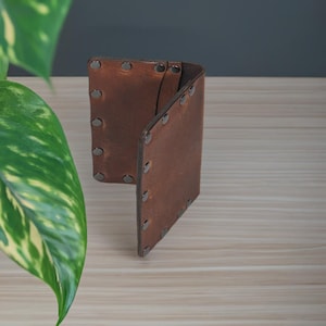 May include: A brown leather wallet with dark metal rivets along the edges. The wallet is open, revealing the interior. The leather has a slightly distressed appearance. The wallet is on a light wood surface.