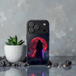 May include: Black phone case featuring a dark figure with a red and purple lightsaber against a red moon backdrop. The case has a glossy finish and a black camera module. A small succulent plant in a white pot is to the left.