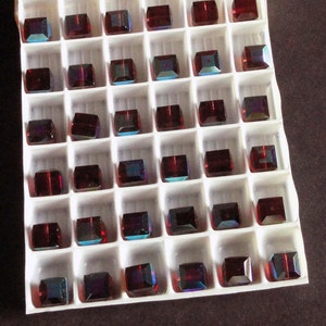 May include: A white plastic tray with 48 square compartments, each containing a dark red square glass bead. The beads have a slight iridescent sheen.