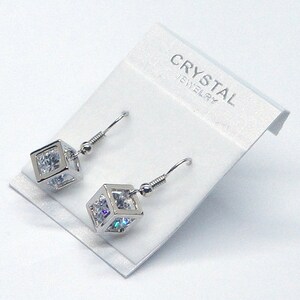May include: A pair of silver dangle earrings with a cubic zirconia stone set in a cube shape. The earrings are on a white card with the text "CRYSTAL JEWELRY".