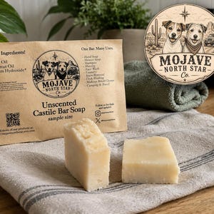 May include: A tan paper pouch labeled Unscented Castile Bar Soap by Mojave North Star Co. with a list of ingredients and uses. Two rectangular off-white soap bars sit in front of the pouch. The logo features two dogs, a cactus, and a vehicle.