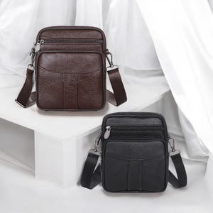 May include: Two leather crossbody bags. One is dark brown, the other is black. Both have a rectangular shape with a front pocket, a top zipper, and an adjustable shoulder strap. The bags are displayed against a white background.