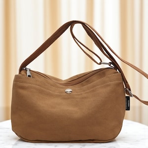 以下が含まれることがあります： ジッパー開閉と調節可能なストラップが付いた、茶色のキャンバスのクロスボディバッグ。 バッグは丸みを帯びた形で、前面に小さな金属製のボタンが付いています。 ストラップはバッグ自体よりも濃い茶色です。