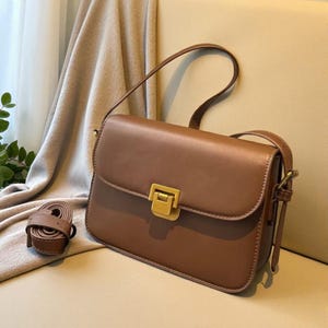 Può includere: Una borsa in pelle marrone con chiusura dorata. La borsa ha una forma rettangolare con chiusura a patta e tracolla. Una cinghia marrone arrotolata è accanto alla borsa.