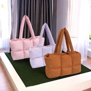 Puede incluir: Tres bolsos tote acolchados en colores pastel: rosa, lavanda y marrón. Cada bolso tiene forma rectangular con un diseño acolchado y asas acolchadas a juego. Los bolsos se exhiben sobre una superficie blanca con una alfombra verde.