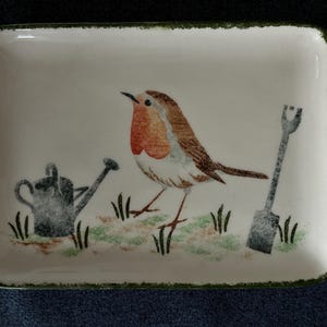 May include: A rectangular ceramic tray with a cream-coloured base and a dark green speckled rim. The centre features a detailed illustration of a robin with a red breast, positioned between a grey watering can and a grey garden shovel.