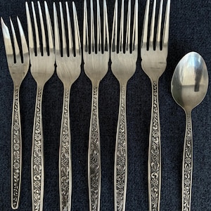 May include: A set of silver-coloured forks and a spoon. The cutlery has a floral design on the handles. The forks are arranged in a row, with the spoon to the right. The background is a dark blue.