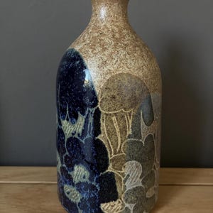 May include: A ceramic bottle with a cork stopper featuring a speckled beige neck and a body decorated with a dark blue and tan floral pattern. The surface has a textured finish with visible etched outlines of flower shapes across the lower section.