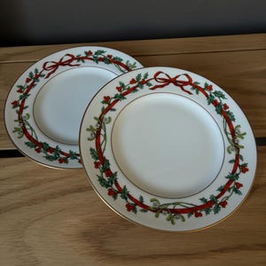 May include: Two white ceramic plates with a gold rim, each featuring a decorative border pattern of red ribbons, green holly leaves, and red berries arranged in a circular wreath design around the centre of the plate.