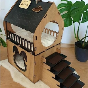 May include: Wooden pet house with a house-shaped design, featuring a black roof, a bunny silhouette, and a set of stairs. The pet house has a light-colored wooden frame and a white fluffy bed inside.