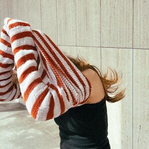 Könnte beinhalten: Ein Paar lange, gestreifte Armstulpen in Rot und Weiß. Die Armstulpen sind aus Strickmaterial und werden hochgehalten. Die Person trägt ein schwarzes Oberteil. Der Hintergrund ist eine hellfarbige Wand.