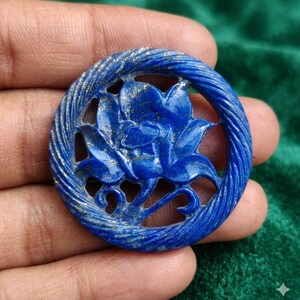 May include: A circular lapis lazuli pendant featuring a carved lotus flower design in the center. The outer edge of the pendant has a textured, twisted rope pattern. The stone is a deep blue color with visible gold-colored pyrite flecks throughout.