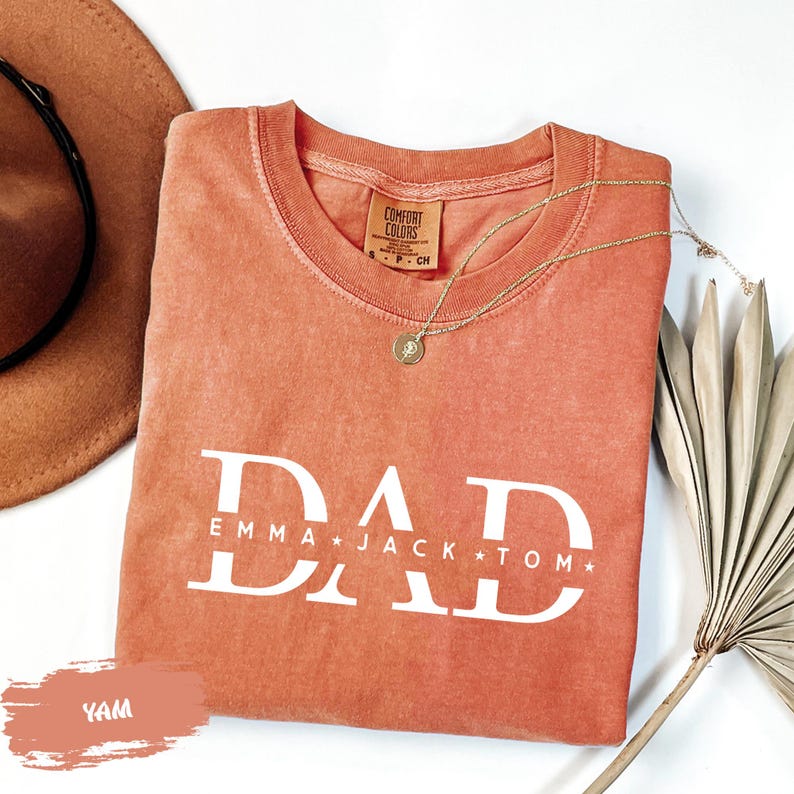 a red t-shirt with the text "DAD" printed on it, along with the names "Emma Jack Tom" below it. The t-shirt is laid out on a white surface, accompanied by a brown hat, a necklace, and a plant.