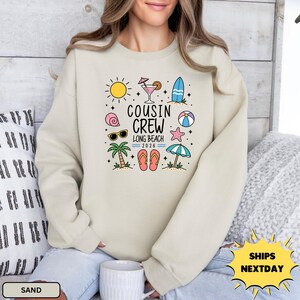 May include: A sand-colored crewneck sweatshirt featuring a graphic print with the text "COUSIN CREW LONG BEACH 2026" surrounded by colorful summer icons including a sun, cocktail, surfboard, seashell, sunglasses, palm tree, flip-flops, and beach umbrella.