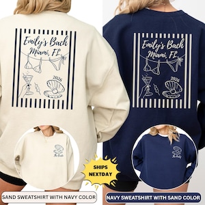 May include: Two sweatshirts, one sand-colored and one navy, featuring a graphic design for a bachelorette party. The design includes the text "Emily's Bach Miami, FL 2026" with bikini tops, a cocktail, and a seashell. The sand sweatshirt has a small seashell graphic that says "The Bride".