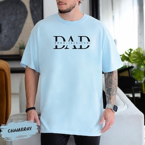 A man wearing a light blue t-shirt with the text "DAD" printed on it, standing in a room with a plant in the background.