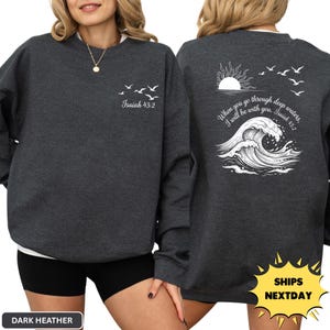 May include: A dark heather gray crewneck sweatshirt featuring a white graphic print. The front displays small birds and the text Isaiah 43:2. The back features a large sun, birds, a crashing ocean wave, and the text When you go through deep waters, I will be with you. Isaiah 43:2.