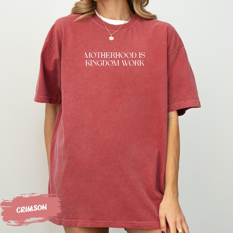 A person wearing a red t-shirt with the text "MOTHERHOOD IS KINGDOM WORK" printed on it.
