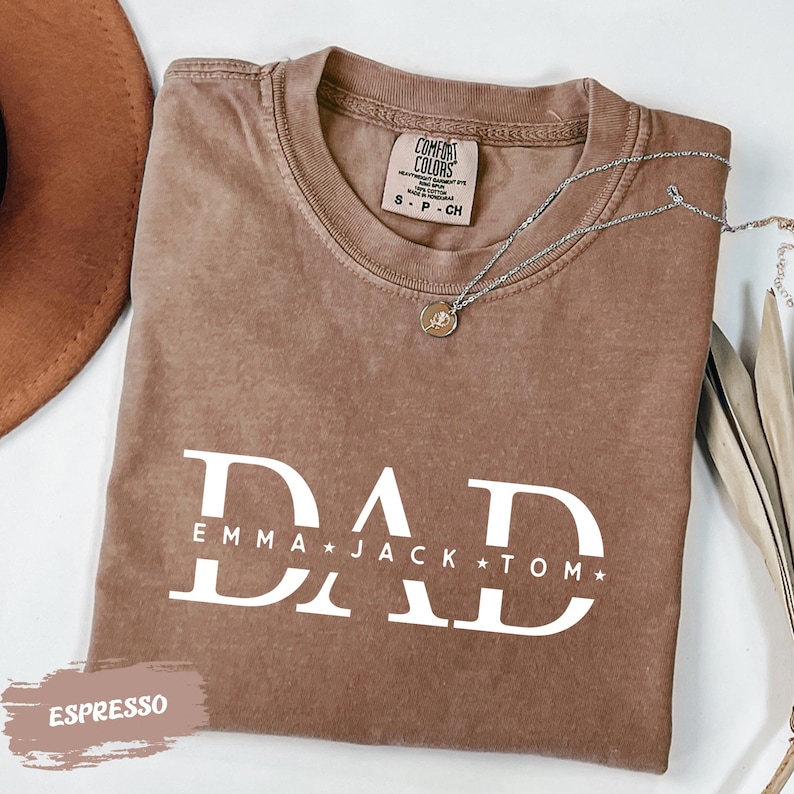 A brown t-shirt with the text "DAD" printed on it, along with the names "Emma Jack Tom" below it. The t-shirt is laid out on a white surface, and there is a necklace and a hat nearby.