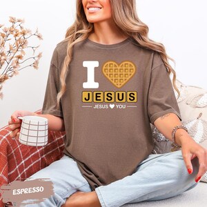 A woman sitting on a couch, wearing a brown t-shirt with a heart-shaped waffle and text that says "I ❤️ JESUS ❤️ YOU". She is holding a coffee mug and appears to be enjoying a relaxed moment.