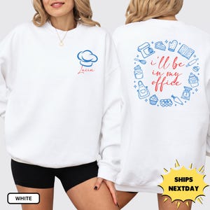 May include: A white crewneck sweatshirt featuring a blue chef hat icon and the name Lucia in red on the front left chest. The back displays a circular arrangement of blue baking icons surrounding the red text I'll be in my office.